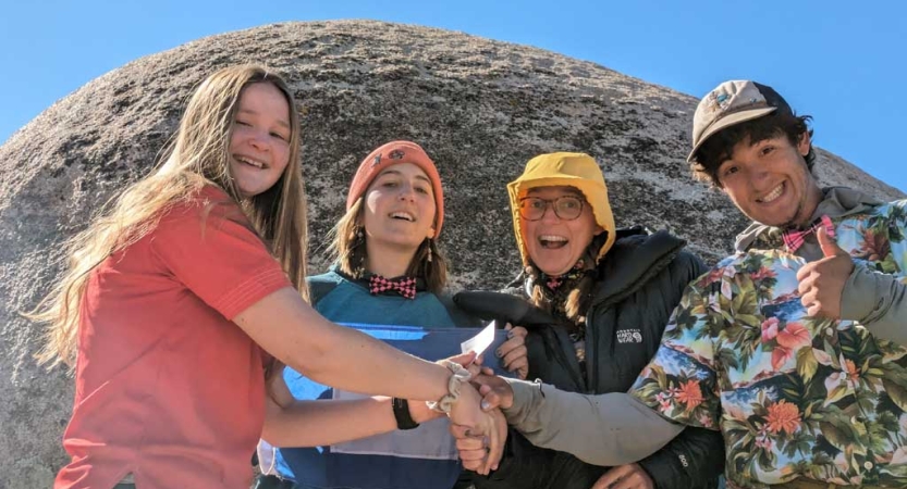 Four people grasp hands and smile while standing in front of a large boulder.